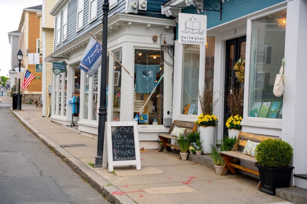 A quaint street scene shows the Saltwater Bookstore with a chalkboard sign that reads, “It’s a good day for a new book!” For those wondering where to find independent bookstores in Massachusetts, this charming spot is not to be missed. Benches and yellow flowers line the storefront.