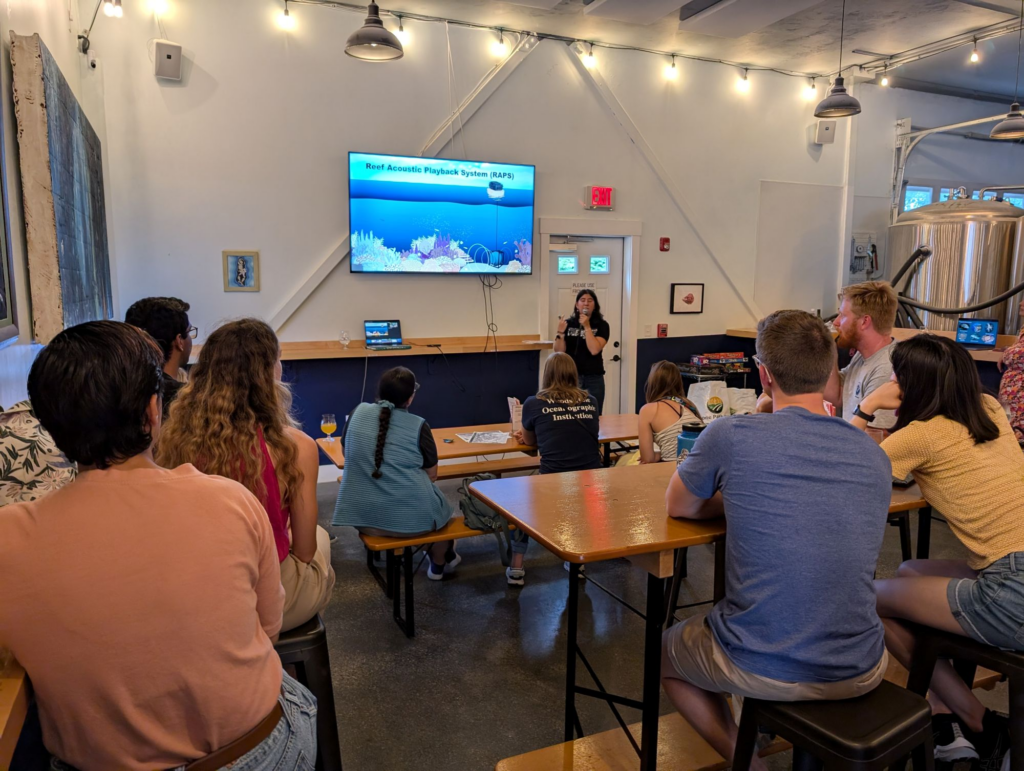 Patrons sit at wooden tables watching a sicence presentation.