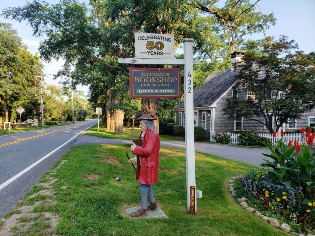 A statue of a revolutionary war-era person underneath a wooden sign that reads "Titcomb's bookshop" on the side of a paved roadway.