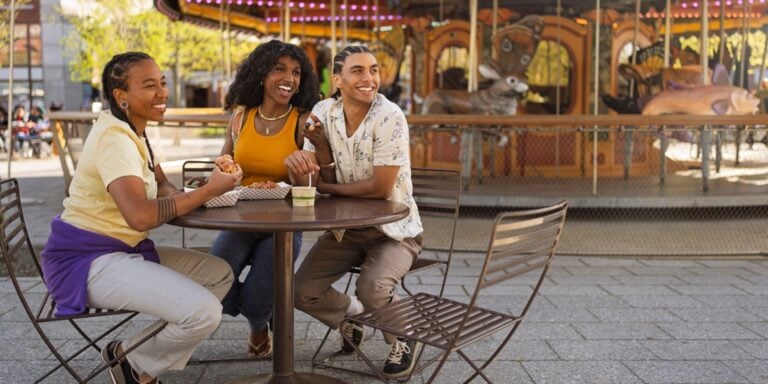 Three friends sit at an outdoor table, smiling and enjoying food and drinks near a carousel during Pride in Massachusetts. The scene is bright and cheerful, with colorful lights and carousel animals in the background.