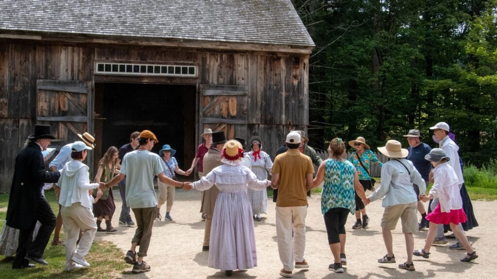 A group of people holding hands in a circle, dancing outdoors in front of a rustic wooden barn; many wear hats and light summer clothes, evoking history at a lively community event where everyone enjoys extra time together on a sunny day.