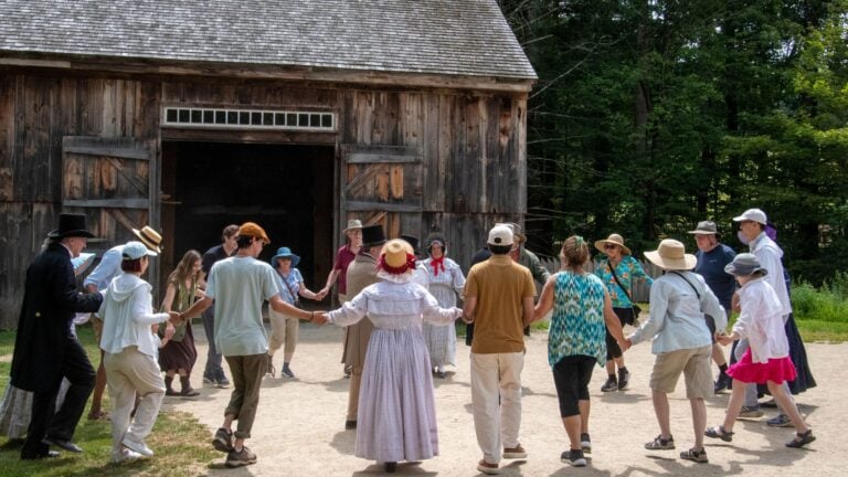 A group of people holding hands in a circle, dancing outdoors in front of a rustic wooden barn; many wear hats and light summer clothes, evoking history at a lively community event where everyone enjoys extra time together on a sunny day.
