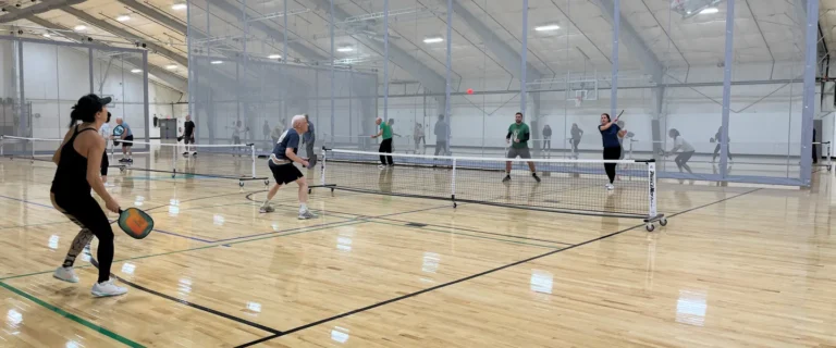 People play pickleball in a large indoor sports facility with polished wooden floors, high ceilings, and multiple courts divided by nets and barriers. Pickleball players of various ages are actively engaged in matches.