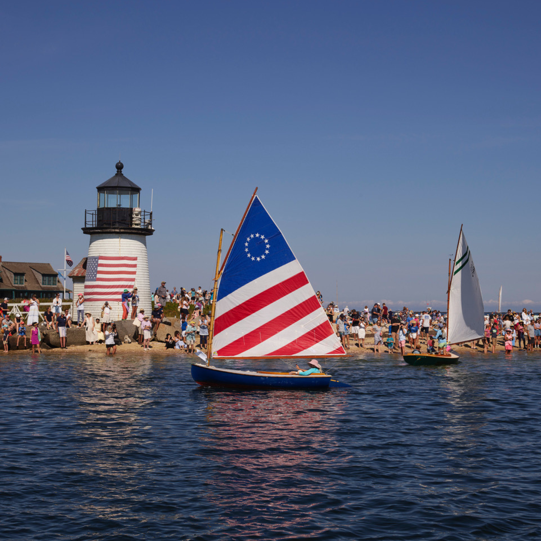 A boat with an American flag sail glides in front of a lighthouse and the coast.