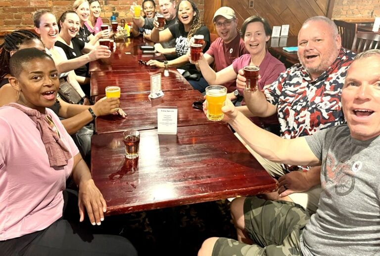 A group of people sit around a wooden table in a restaurant, smiling and raising glasses of craft beer and other drinks in a toast. The atmosphere appears cheerful and celebratory.