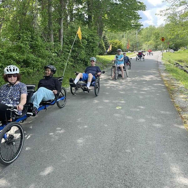 A group of people ride recumbent tricycles on a paved path surrounded by greenery, wearing helmets and enjoying a sunny day. Flags are attached to some of the trikes for visibility—a great scene for any travel guide seeking unique outdoor experiences.