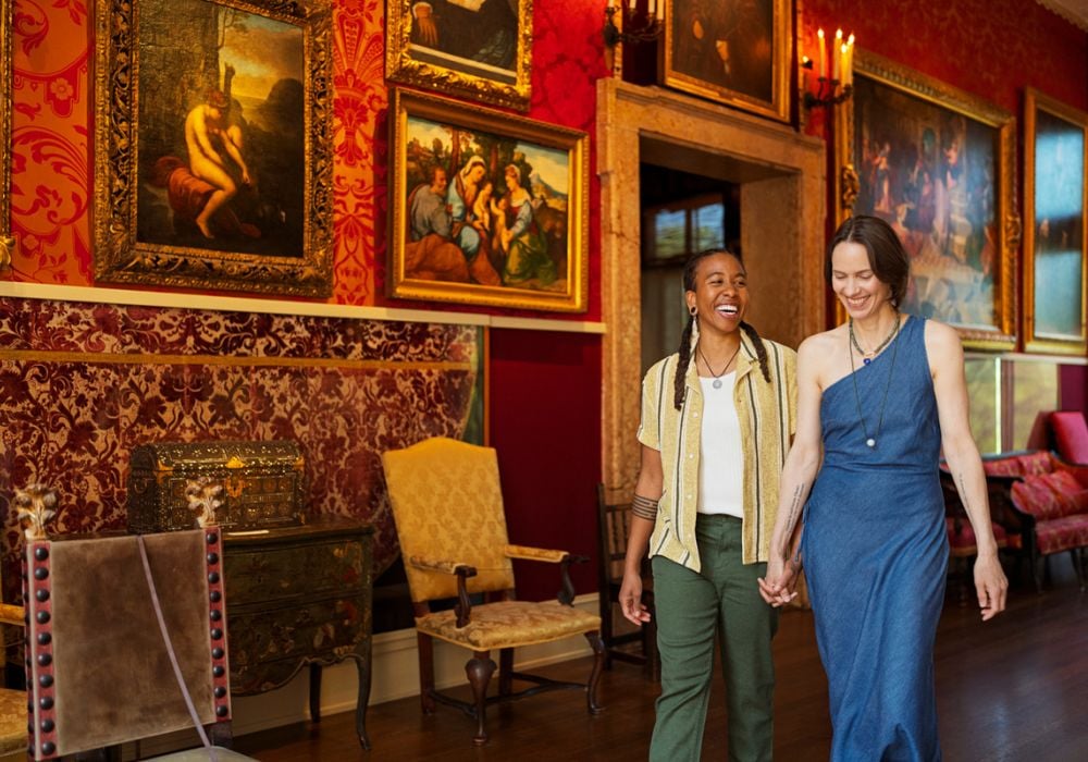 Two people hold hands and smile while walking through an ornate Massachusetts art gallery with red wallpaper, gold-framed paintings, and antique furniture—an inviting scene for LGBTQ+ travel and tourism.