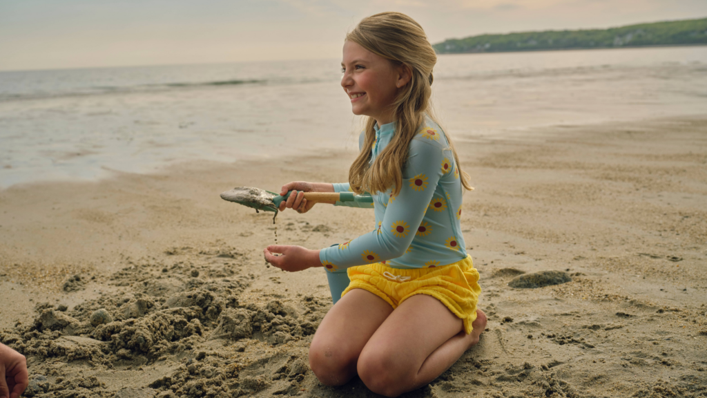 A young girl in a blue long-sleeve shirt with yellow flowers and yellow shorts kneels on the coast, smiling as she holds a small fish in her hands near the shoreline, enjoying some extra time by the water.