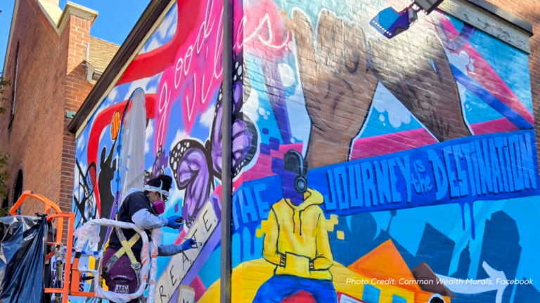 An artist in protective gear paints a colorful mural on a building wall, featuring raised fists, butterflies, and bold text reading THE JOURNEY IS THE DESTINATION. Bright colors and patterns fill the mural.
