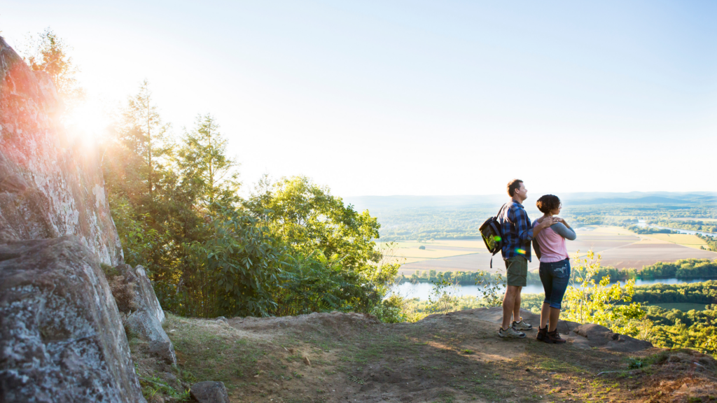 A man and woman stand on a rocky hilltop looking out over a scenic view of fields, trees, and a river, with the sun shining through the trees on the left. The man has a backpack, and the weather appears clear.