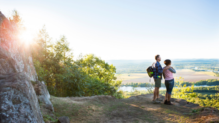 A man and woman stand on a rocky hilltop looking out over a scenic view of fields, trees, and a river, with the sun shining through the trees on the left. The man has a backpack, and the weather appears clear.