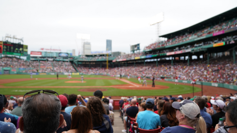 A large crowd enjoys a Massachusetts Sports baseball game at a stadium, with players on the field and city buildings visible in the background under a cloudy sky.