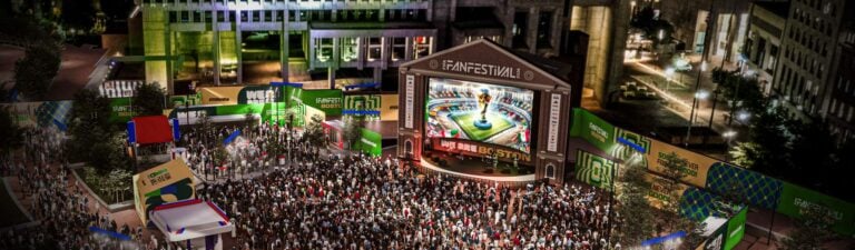 A large crowd gathers at an outdoor festival in front of a stage with a big screen showing a soccer celebration. Bright lights, colorful banners, and city buildings surround the lively summer of soccer event.