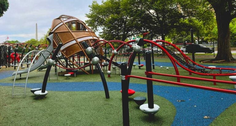 Summertime Means Playground Time at this modern spot with climbing structures, slides, and curved red bars on soft green and blue rubber flooring, all surrounded by trees and a black metal fence.