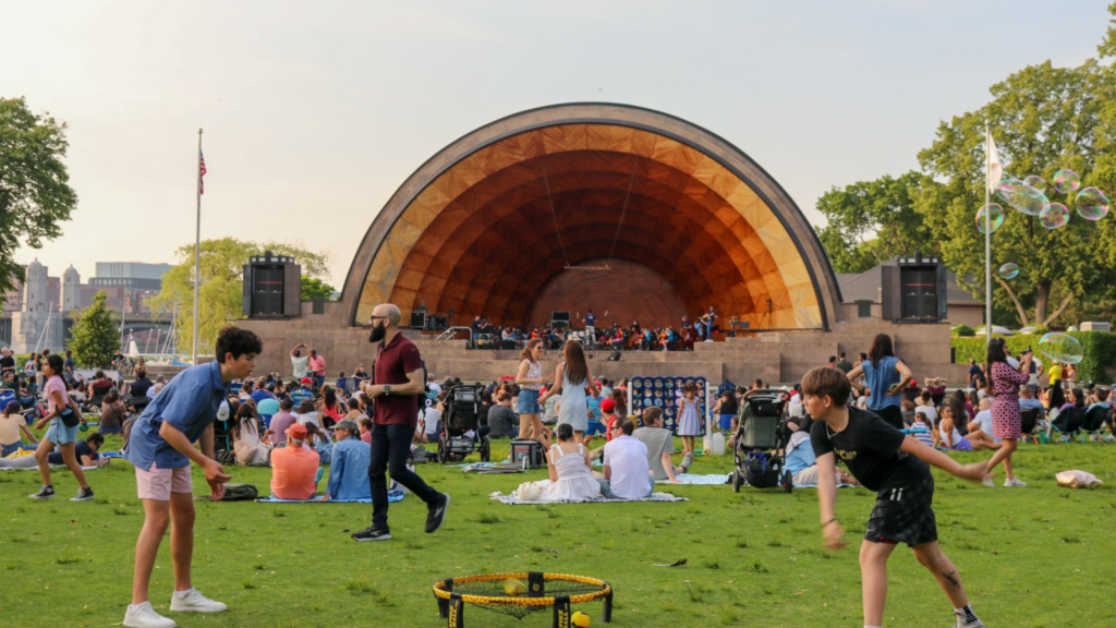 People enjoy a sunny day at an outdoor concert in front of a large wooden bandshell. Children play on the grass, and groups sit on blankets, watching musicians perform on stage. Trees and bubbles are visible in the background.
