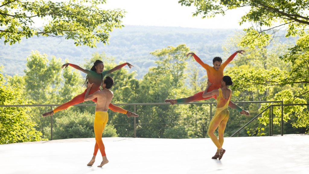 Four ballet dancers in colorful costumes perform outdoors on a stage surrounded by lush green trees, with two dancers lifting their partners against a scenic, sunlit backdrop of hills and sky.