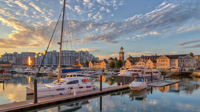 A marina at sunset with sailboats and yachts docked along wooden piers; waterfront buildings, a lighthouse, and modern apartments set the scene for a perfect Greater Boston road trip under a partly cloudy sky.