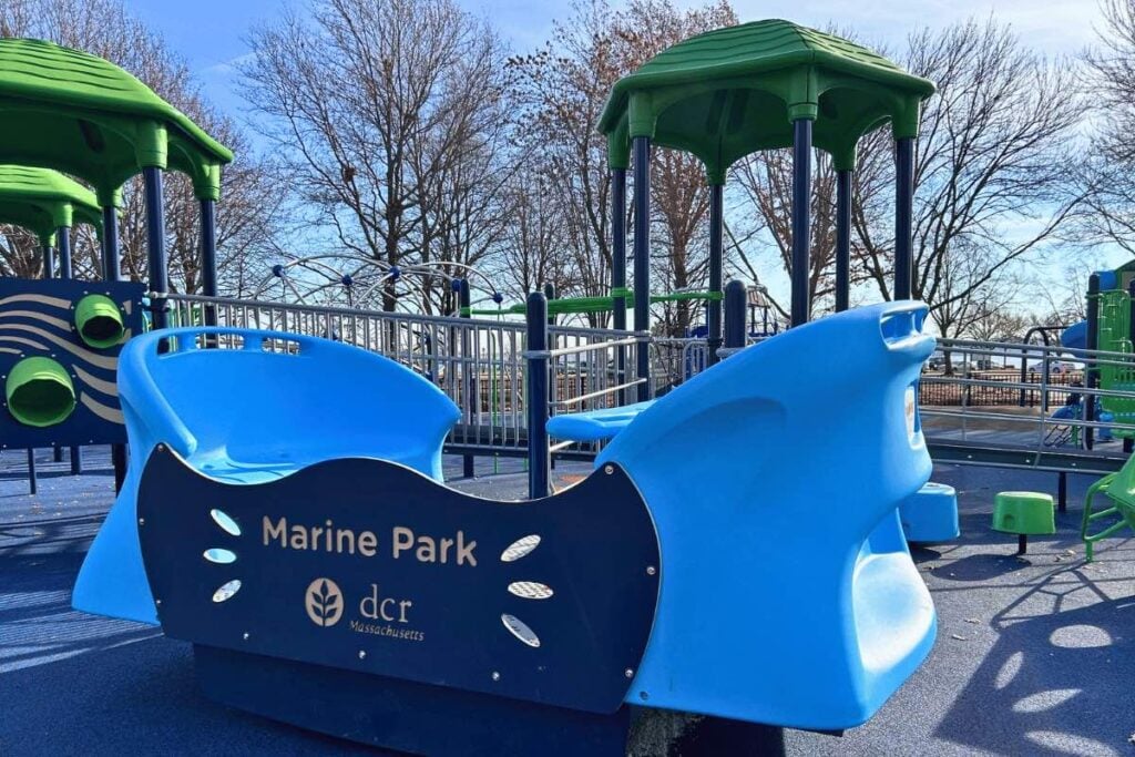 A blue play structure labeled Marine Park with dcr Massachusetts sits on a playground with green equipment and trees without leaves in the background.