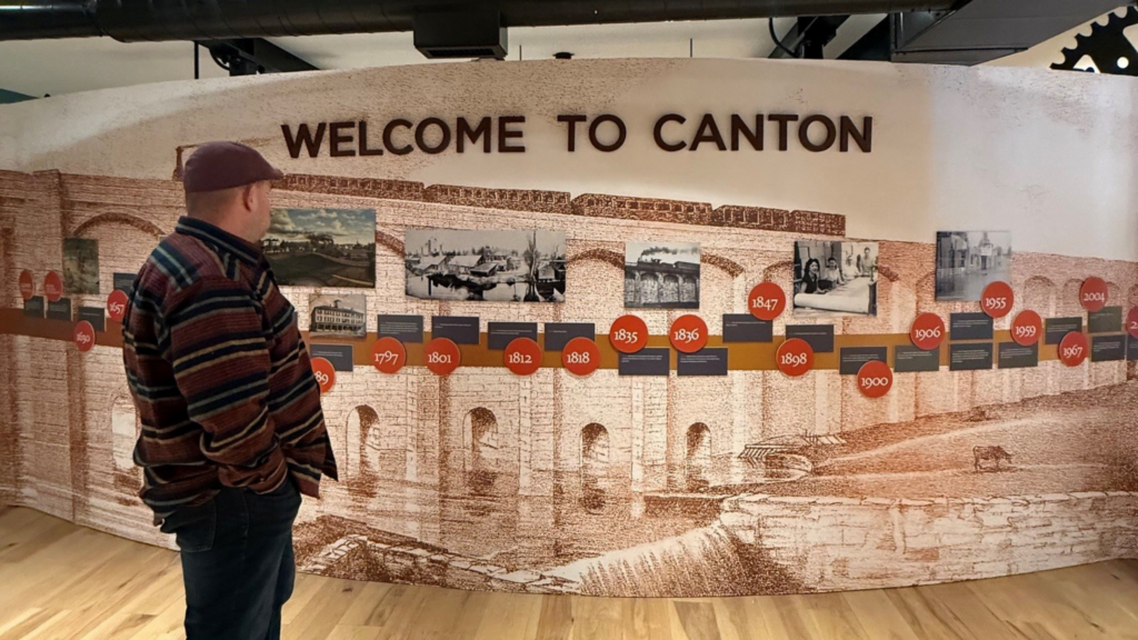A man in a striped shirt and maroon hat stands in front of a large display titled WELCOME TO CANTON, featuring a historical timeline with images and dates—an ideal stop for any Greater Boston road trip.