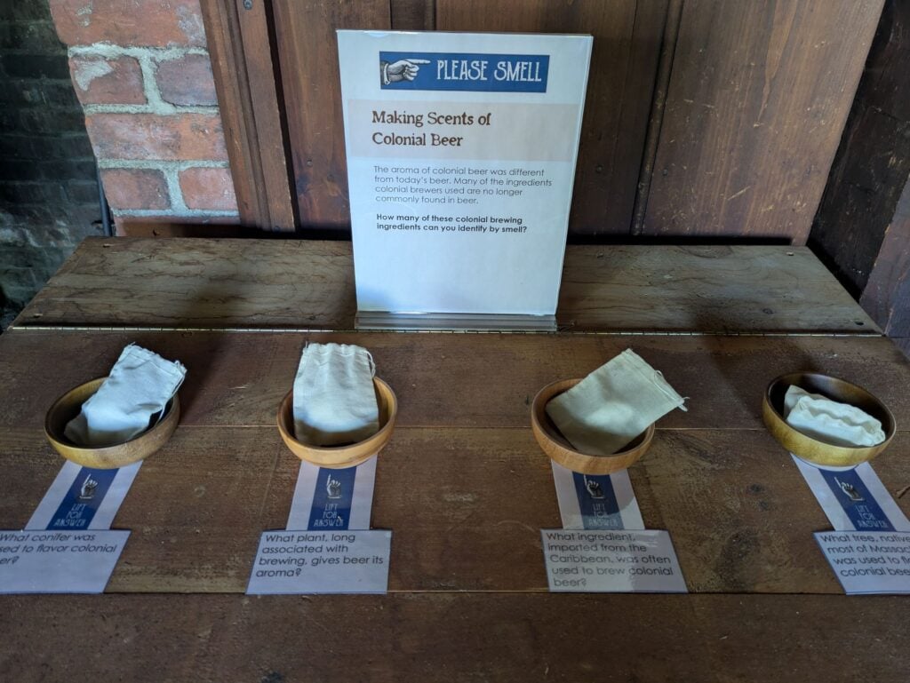 Four wooden bowls with cloth squares are arranged on a wooden table beneath a sign about the scents of colonial beer, each bowl labeled with questions about beer ingredients and aromas.