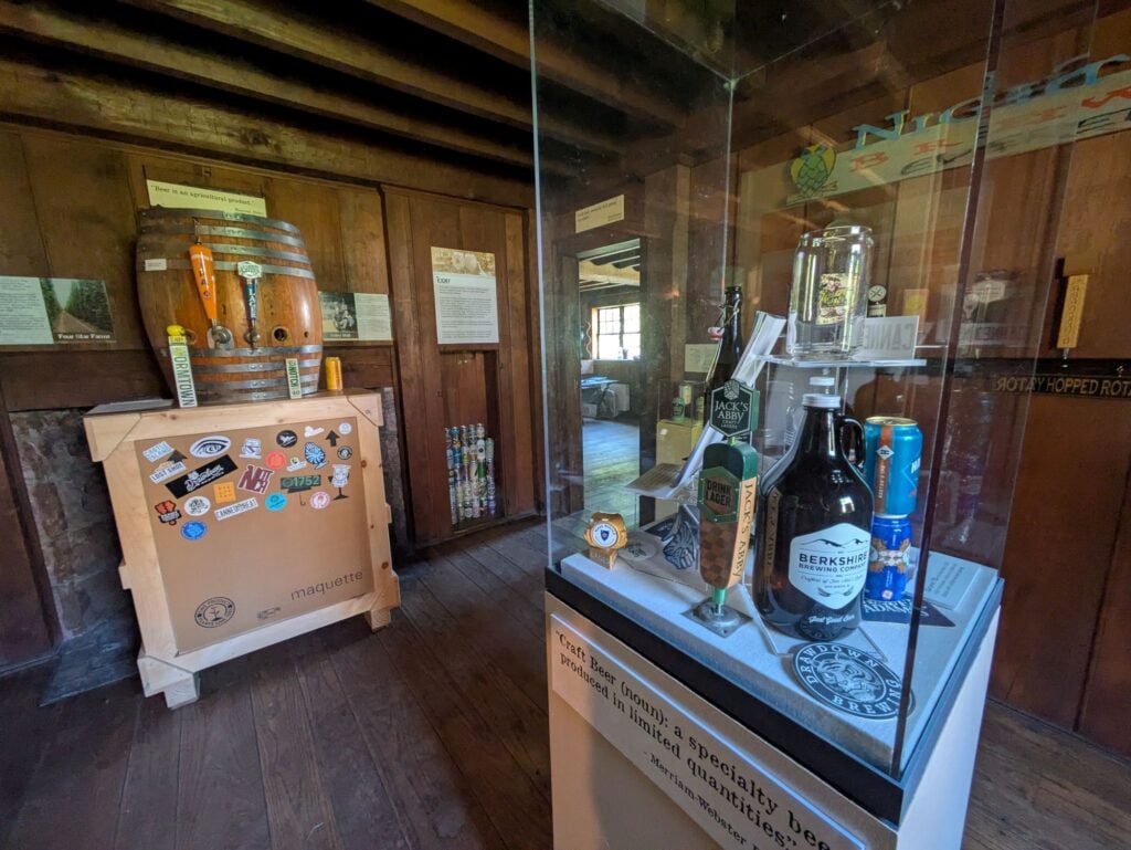 A museum exhibit displays various bottled beverages and glassware in a glass case. Nearby, a large barrel sits on a box decorated with stickers. The room has wooden walls, floors, and historical information panels.