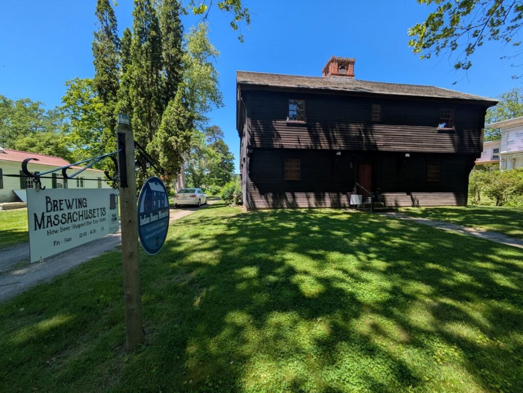 A large, dark wooden house with small windows sits on a grassy lawn. A sign in the foreground reads “Brewing Massachusetts,” hinting at its craft beverage tradition, next to a gravel driveway under a clear blue sky. Trees and other houses are in the background.