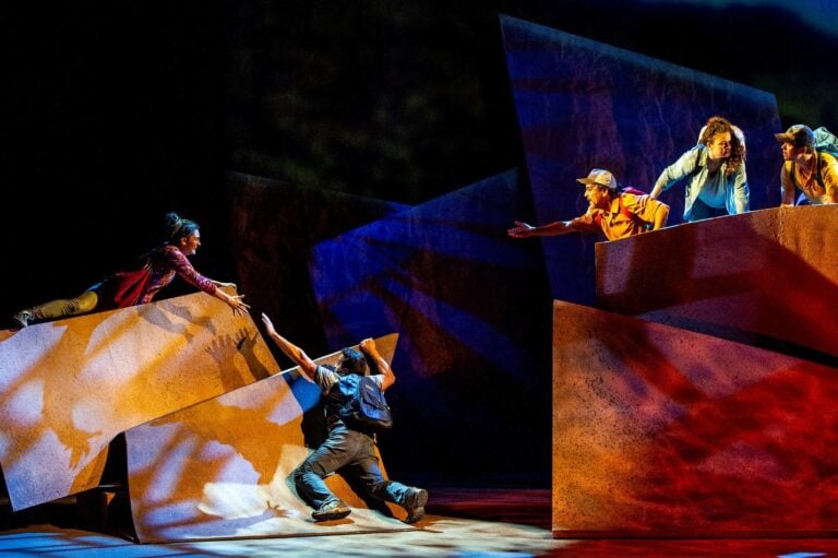 Actors on stage with Independent Theater Companies in Massachusetts perform a dramatic scene, reaching out to each other across large, angular set pieces with colorful lighting, creating a sense of urgency and separation.