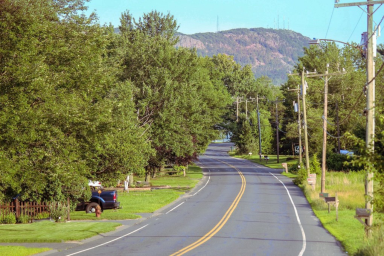 A winding two-lane road, part of The Best Scenic Roads in Massachusetts, runs through a lush, green rural area with trees on both sides, utility poles lining the right, and a distant rocky hill under a clear blue sky.