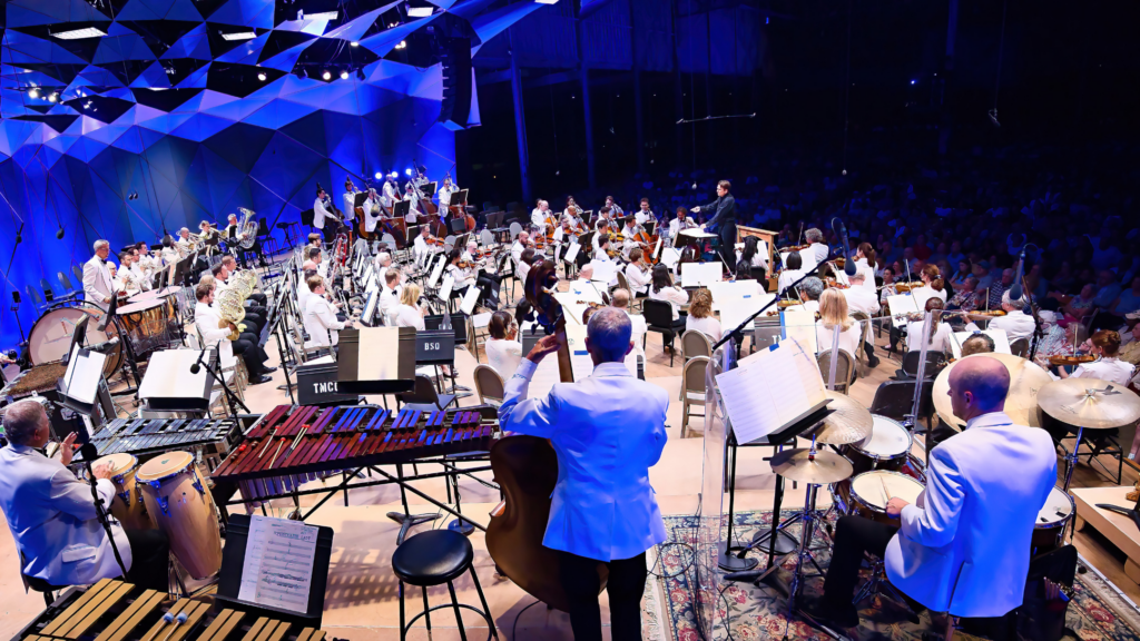 A large orchestra performs on stage in a concert hall, with musicians in white jackets playing various instruments under blue lighting. The conductor stands in front, leading the ensemble, as an audience watches.