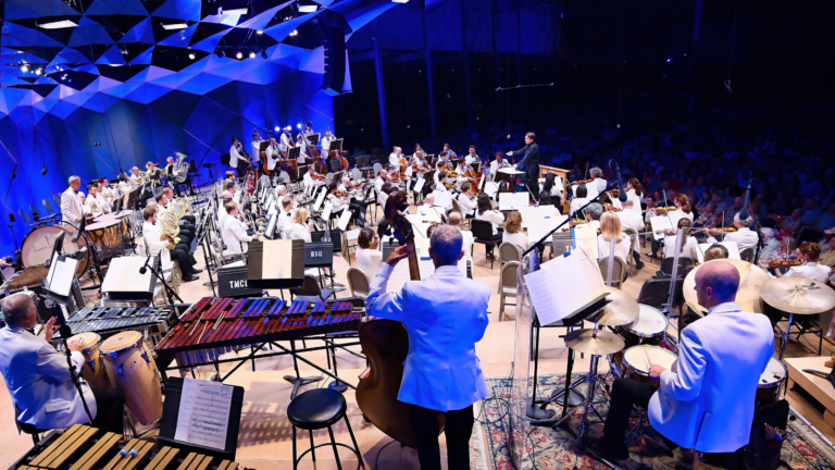 A large orchestra performs on stage in a concert hall, with musicians in white jackets playing various instruments under blue lighting. The conductor stands in front, leading the ensemble, as an audience watches.
