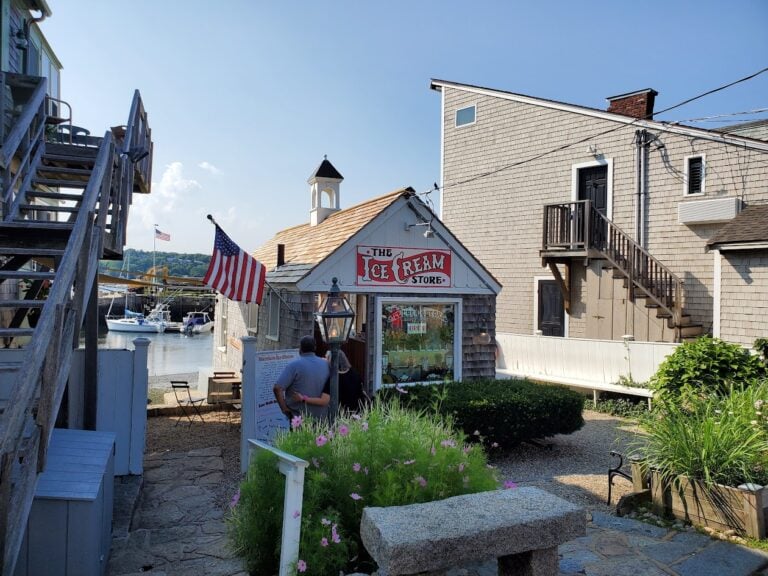 A small ice cream shop with an American flag outside sits by a harbor, surrounded by flowers, wooden buildings, and two people reading a menu. On this clear and sunny day, it’s the perfect stop along the Massachusetts Ice Cream Trail to celebrate National Ice Cream Day.