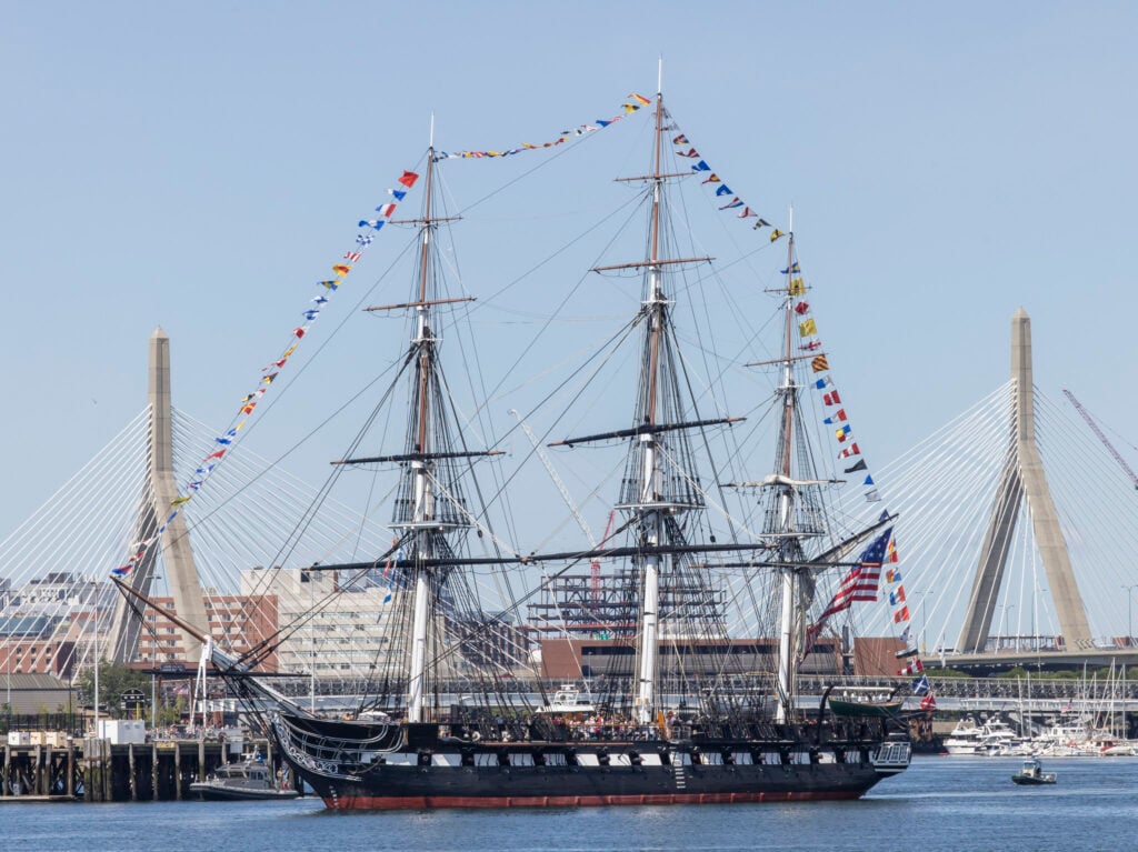 A historic tall ship with three masts, adorned with colorful signal flags, sails on a harbor during Sail Boston, with a cable-stayed bridge and city buildings in the background under a clear blue sky.