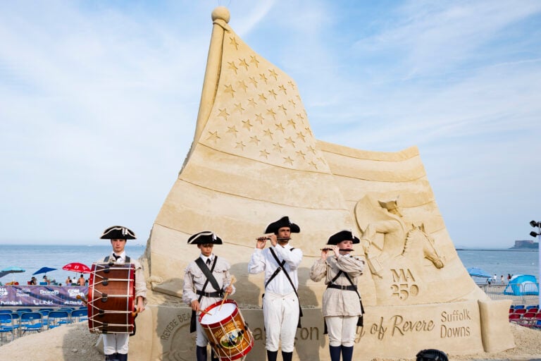 Four people in colonial attire play instruments before a large piece of sand art shaped like a waving American flag at the beach. The sculpture reads MA 250 and Suffolk Downs, with engraved stars and stripes, capturing the magic of sand art in its intricate detail.