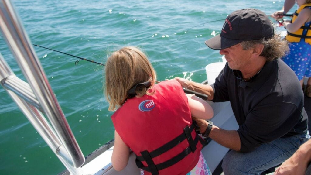 An adult helps a child in a red life jacket fish from a boat on a sunny day. The water is calm and another child in a yellow life jacket is visible in the background.