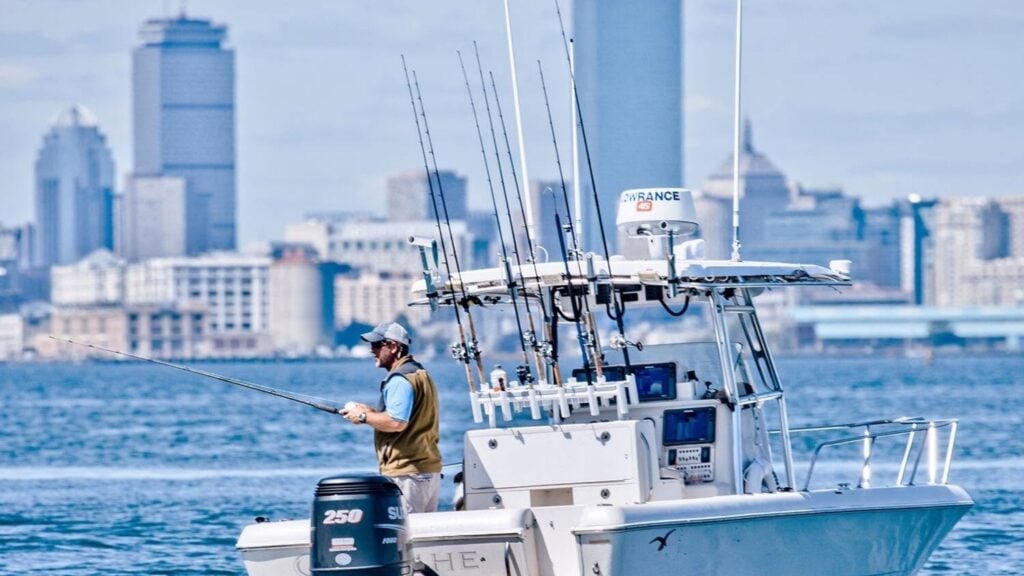 A man enjoys charter fishing from a white motorboat on the water, with several fishing rods upright behind him. A city skyline with tall buildings is visible in the background under a partly cloudy Massachusetts sky.