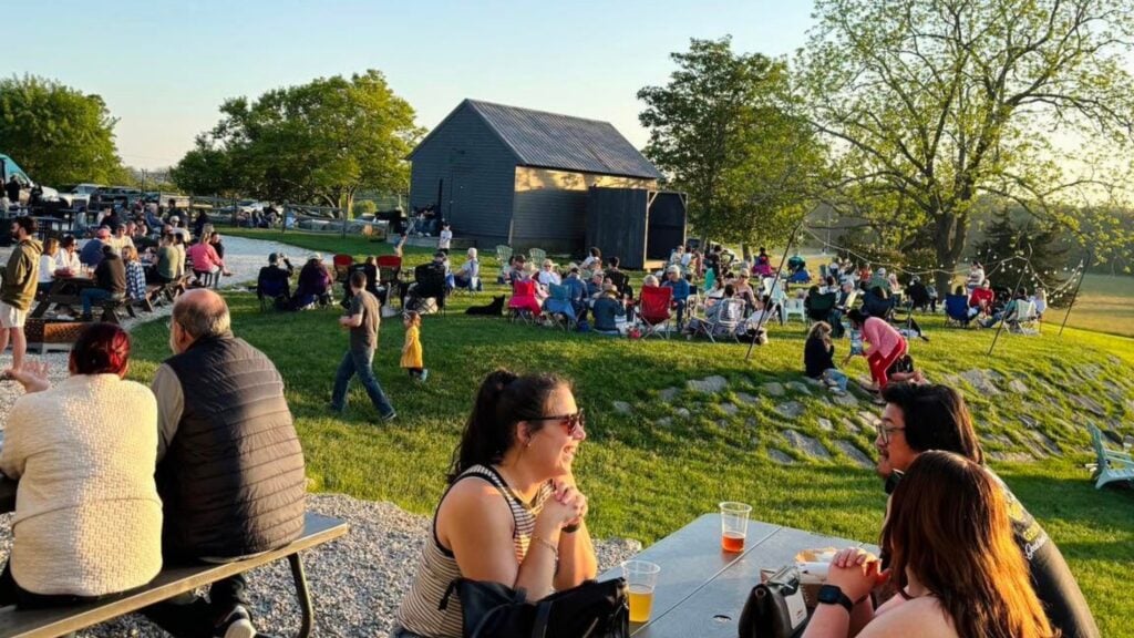 People sit at picnic tables and on lawn chairs outdoors near a barn in Southeastern Massachusetts, enjoying drinks and conversation at a lively community event on a sunny day—the perfect stop to explore during your next road trip.