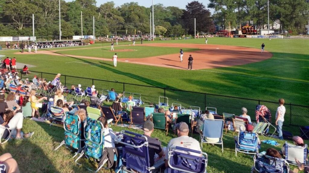 Spectators sit on lawn chairs watching a baseball game on a sunny day. Players are on the field, and trees line the background. The scoreboard and dugouts are visible in the distance.