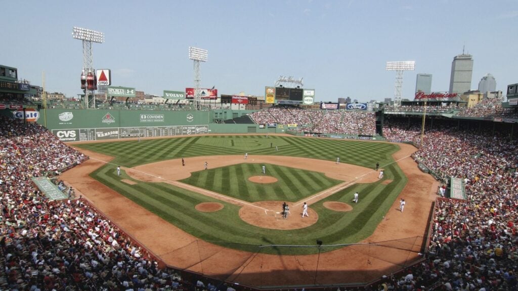 A wide view of a baseball game at Fenway Park with players on the field showcasing baseball in action, fans filling the stands, and the Boston city skyline in the background under a clear Massachusetts sky.