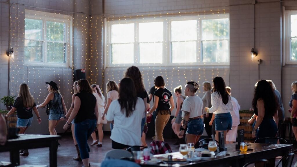 A group of people enjoy line dancing together in a sunlit indoor space with large windows and string lights. Some tables with drinks and plates are visible in the foreground, adding to the lively atmosphere typical of gatherings in Massachusetts.