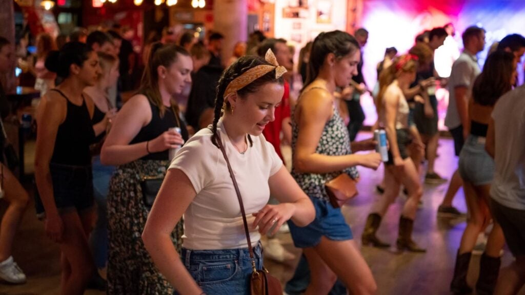 A group of people enjoys line dancing indoors at a lively event. Most wear casual summer clothing, and some hold drinks. The background is softly lit with colorful lights, creating a festive atmosphere reminiscent of celebrations in Massachusetts.
