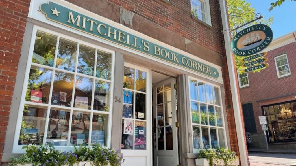 A brick bookstore with large front windows and a green sign reading Mitchells Book Corner. Potted flowers line the windows, making it a charming stop during your Five-Star Weekend or luxury getaway in town. A hanging sign displays the shop’s name above the entrance.