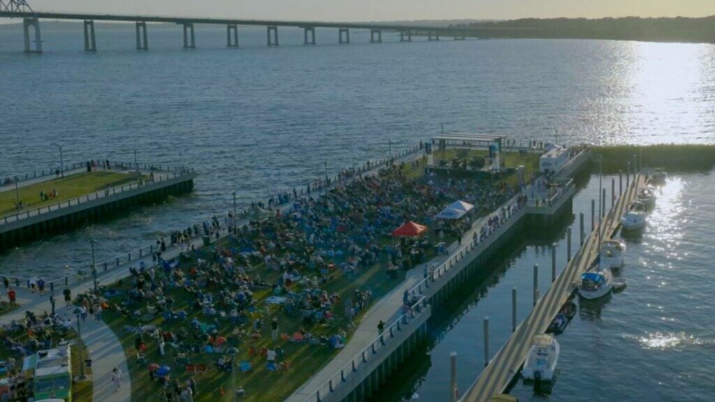 A large crowd gathers on a grassy pier by the water in Southeastern Massachusetts for an outdoor event, with boats docked nearby and a bridge spanning the background under a bright, setting sun—a perfect scene to explore during your next road trip.