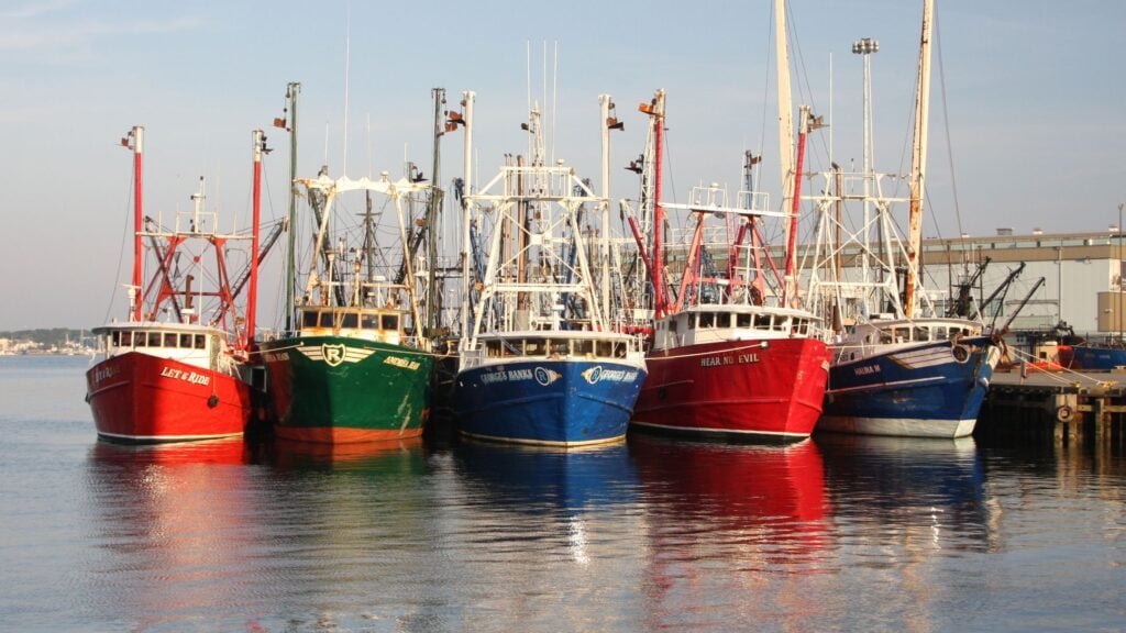 Several colorful fishing boats, mainly red and blue, are docked side by side at a harbor on a calm, sunny day in Southeastern Massachusetts, with industrial buildings visible in the background—a perfect spot to explore on your next road trip.