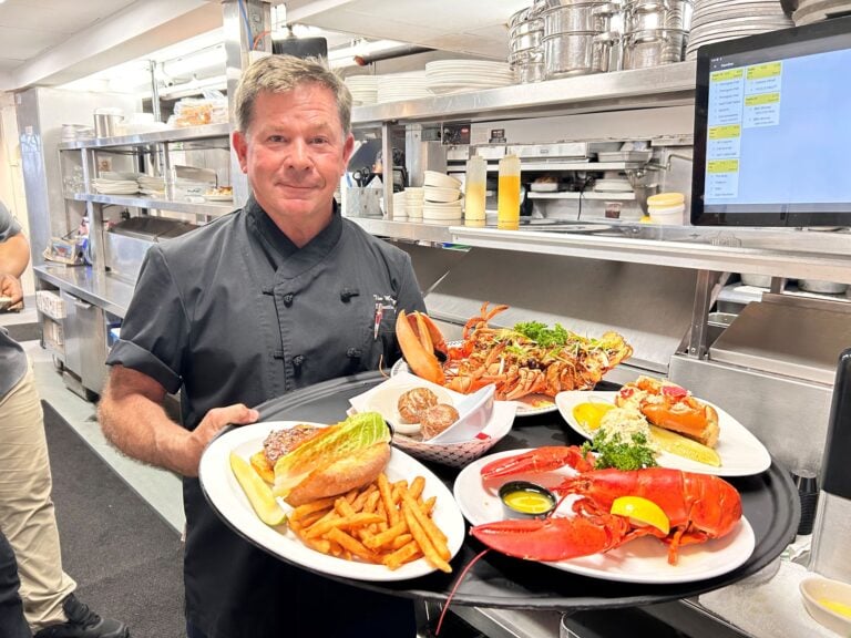 A chef in a kitchen holds a large tray with plates of lobster, fries, a sandwich, and other seafood dishes.