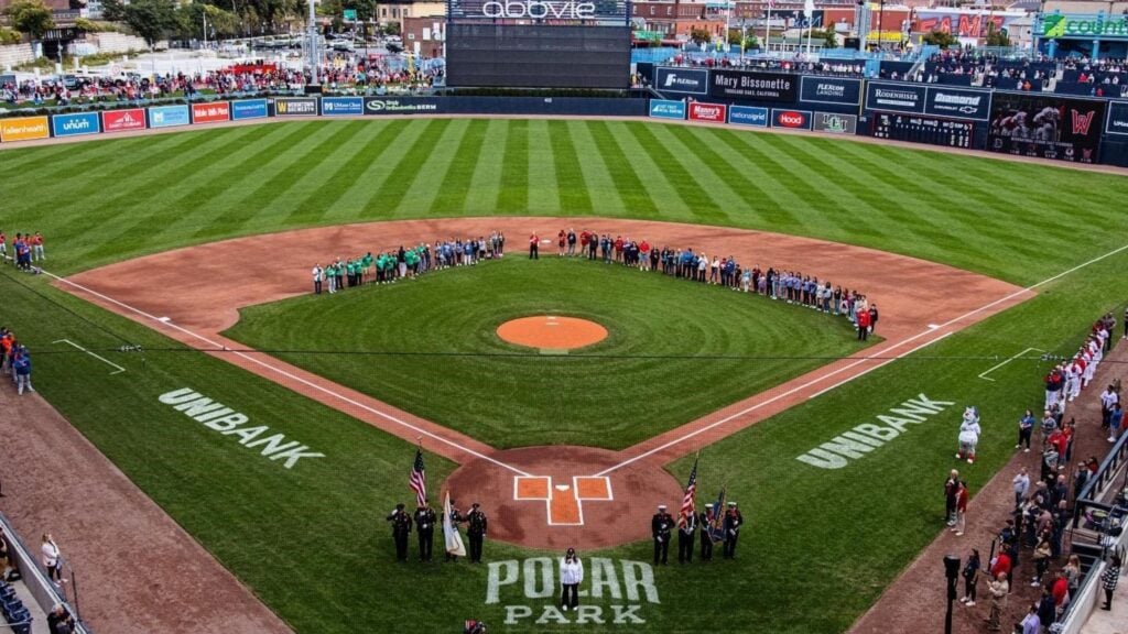 Aerial view of a baseball field in Massachusetts with teams and officials lined up along the diamond, forming a semi-circle. The stadium stands are filled with spectators, and UNIBANK is written on the grass near first and third base.