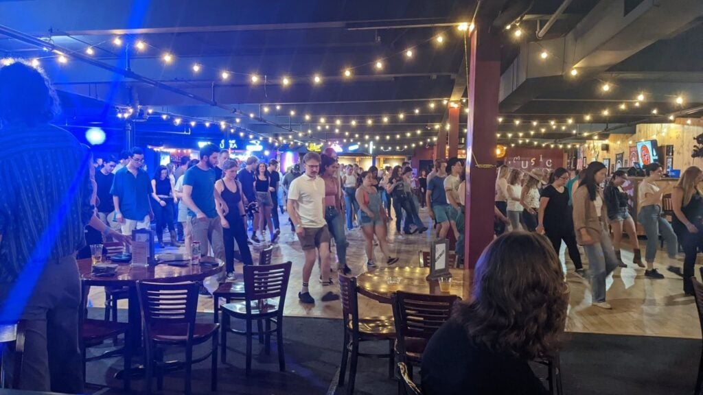 A large group of people dance in a lively, string-lit indoor venue, reminiscent of the best Line Dancing venues Massachusetts has to offer. Some sit at tables in the foreground, while others move energetically on the dance floor. The atmosphere is festive and social.
